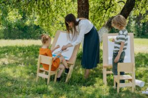 pexels photo 8922967 8922967 A teacher guiding young children in an outdoor painting class under a tree.